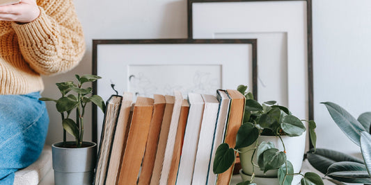 A girl was reading a book on a white colored table along with green plants, properly arranged books, photo frames with a white wall in the background.